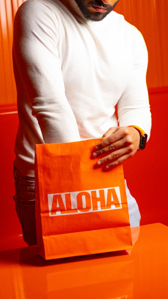 A man stands holding an Aloha branded takeout bag in an orange-themed restaurant setting.